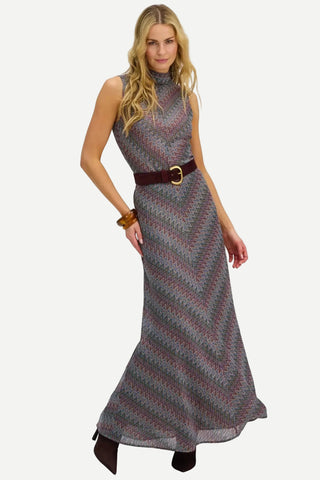 A woman modeling a sleeveless, high-neck maxi dress with a gray, red, and black chevron pattern, styled with a dark brown belt with a gold buckle.