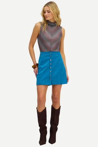 A woman modeling a sleeveless mock-neck top with a geometric pattern in gray, pink, and blue, paired with a bright blue button-front mini skirt and dark brown knee-high boots, against a plain white background.