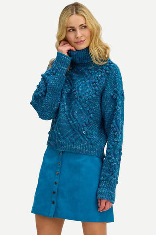 A woman modeling a teal blue cable-knit turtleneck sweater with textured pom-pom details, paired with a matching blue button-front skirt, against a plain white background.